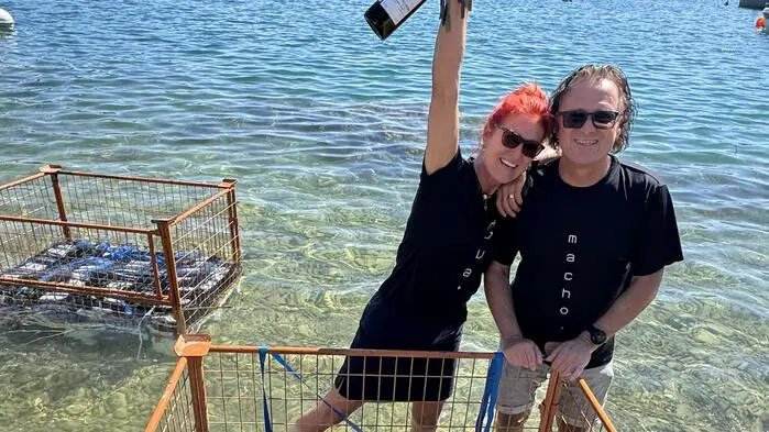 Eine Frau und ein Mann stehen im See hinter einem Metallgerüst, in dem Weinflaschen geschlichtet sind.