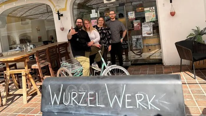 Ein Gruppenfoto, auf dem zwei Männer und zwei Frauen vor einem Restaurant zu sehen sind. Vor ihnen ist eine Tafel mit der Aufschrift „Wurzelwerk“.