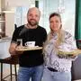 Eine Frau und ein Mann stehen in einem Café und lächeln in die Kamera. Beide halten ein Tablett mit Kaffee in den Händen.