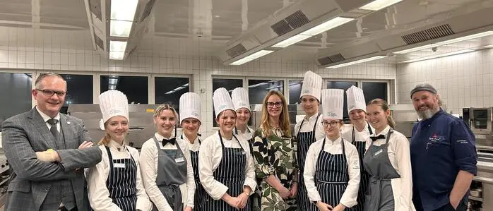 Schülerinnen und Schüler in Koch- und Servicekleidung stehen mit einer Frau und zwei Männern in einer Großküche