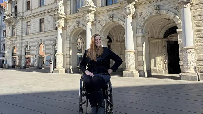 Barbara Sima-Ruml steht mit ihrem Rollstuhl auf dem grazer Hauptplatz und lächelt in Richtung Kamera. Im Hinergrund befindet sich der Eingang zum Rathaus.