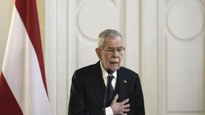 Bundespräsident Alexander Van der Bellen steht auf einem Rednerpult und fasst sich mit der rechten Hand an die Brust. Hinter ihm ist die österreichische Flagge zu sehen