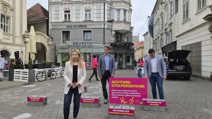 Verena Garber mit weißem Sakko, dunklem Leiberl und Hose sowie hellen Schuhen, steht beim Wahlkampfauftakt an vorderster Stelle; im Hintergrund Philipp Poinntner (Mann mit Sakko und blauem Hemd) und Alexis Pascuttini (Blick nach unten, hellblaues Sakko, weißes Hemd)