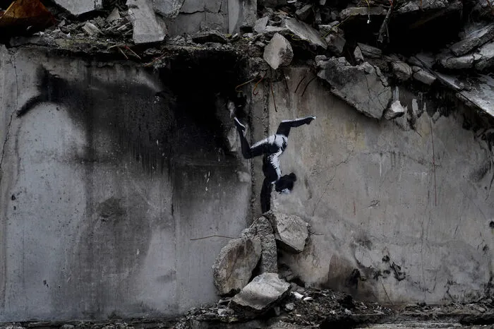 Das Werk zeigt auf der grauen Wand eines kriegszerstörten Hauses ein Mädchen, das scheinbar auf Trümmern einen Handstand macht.