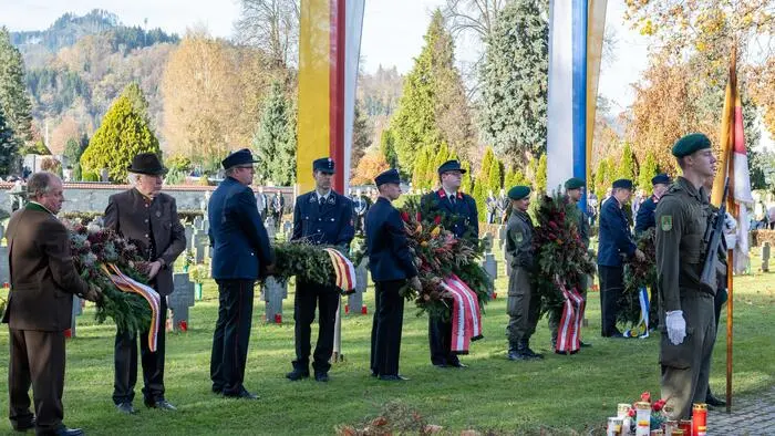 Eine Gruppe uniformierter Personen steht auf einem Friedhof und hält Kränze.