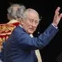 König Charles hebt die Hand zum Gruß. Hinter ihm ist ein Geistlicher zu sehen.