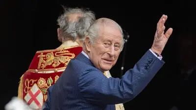 König Charles hebt die Hand zum Gruß. Hinter ihm ist ein Geistlicher zu sehen.