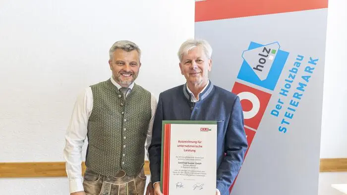 Zwei Männer lächeln in die Kamera. Der Mann rechts hat eine Urkunde in der Hand