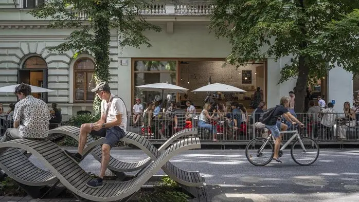 Menschen sitzen in der Kaiserfeldgasse im Gastgarten eines Lokals. Jemand fährt gerade auf dem Rad vorbei. Auf der anderen Straßenseite sitzen Menschen auf öffentlichen Möbeln.