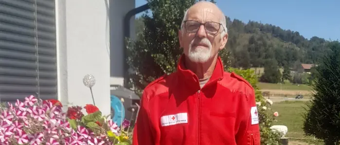 Ein Mann mit Rot-Kreuz-Jacke steht neben Blumen und lacht in die Kamera