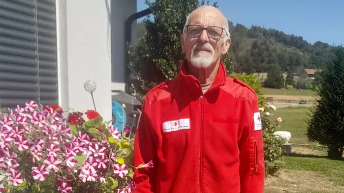 Ein Mann mit Rot-Kreuz-Jacke steht neben Blumen und lacht in die Kamera