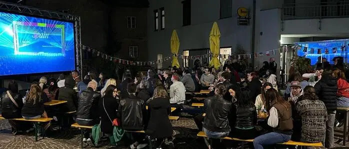 Nächtliche Aufnahme von einer Public-Viewing Veranstaltung; man sieht eine Leinwand und davor sitzen zahlreiche Menschen auf Bierbänken