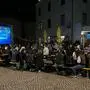 Nächtliche Aufnahme von einer Public-Viewing Veranstaltung; man sieht eine Leinwand und davor sitzen zahlreiche Menschen auf Bierbänken