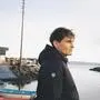 Dunkelhaariger Mann mit Jacke blickt in die Ferne, hinter ihm sind das Meer und Boote zu sehen