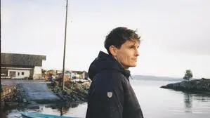Dunkelhaariger Mann mit Jacke blickt in die Ferne, hinter ihm sind das Meer und Boote zu sehen