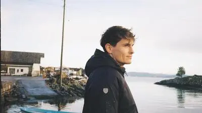 Dunkelhaariger Mann mit Jacke blickt in die Ferne, hinter ihm sind das Meer und Boote zu sehen