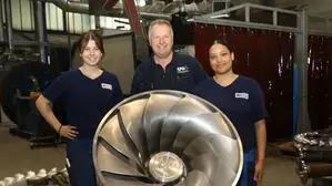 Zwei junge Damen und ein älterer Herr stehen hinter einer Turbine, die in Wasserkraftwerken zum Einsatz kommt.