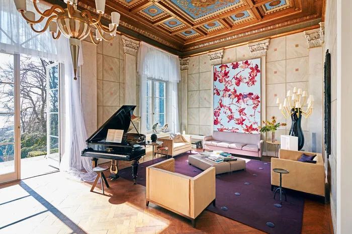 Das Wohnzimmer umfasst eine Wohnfläche von 80 Quadratmetern und erstreckt sich über die gesamte Breite des Hauses. Mit einer Raumhöhe von bis zu sechs Metern sowie der mit Blattgold verzierten Kassettendecke erinnert dieser Raum an einen Saal. Bodentiefe Rundbogenfenster ermöglichen den uneingeschränkten Elbblick und eine direkte Verbindung zum Garten und der Terrasse.