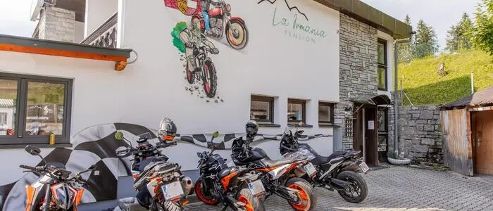 Mehrere Motorräder stehen vor einem hotelähnlichen Gebäude, auf dem zwei Motorräder gezeichnet sind und das Logo der Pension ersichtlich ist