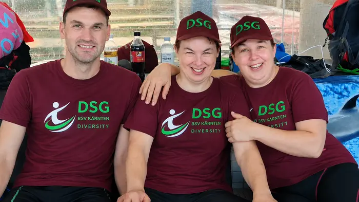 Drei Personen sitzen nebeneinander und tragen die gleiche dunkelrote Uniform mit grüner Schrift und Kappe 