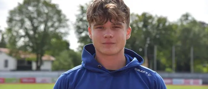 Ein junger Mann mit blauem Oberteil steht auf einer Sportanlage und blickt in die Kamera