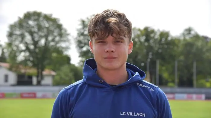 Ein junger Mann mit blauem Oberteil steht auf einer Sportanlage und blickt in die Kamera