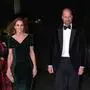 Auf dem Foto schreiten der Prinz und die Prinzessin von Wales über den roten Teppich. Die Prinzessin trägt ein bodenlanges, dunkelgrünes Samtkleid mit leicht drapiertem Ausschnitt und kurzen Ärmeln. Ihr Haar fällt in weichen Wellen über die Schultern, und sie trägt auffällige, lange Ohrringe sowie ein funkelndes Armband und eine Clutch. Prinz William an ihrer Seite trägt einen klassischen Abendlook: schwarzes Samtjackett, weiße Hemdbrust, schwarze Fliege und dunkle Anzughose. Beide wirken gut gelaunt und lächeln, während sie gemeinsam in Richtung Kamera gehen. | Bei der Royal Variety Performance zeigten sich  der Prinz und die Prinzessin im Partnerlook.
