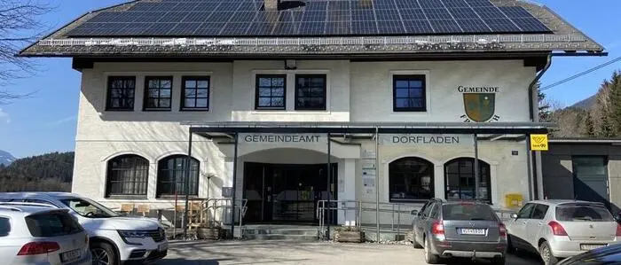 Blick auf ein zweistöckiges Gebäude mit der Aufschrift „Gemeindeamt“ und „Dorfladen“, davor ein Parkplatz mit einigen Autos