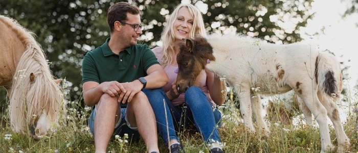Ein Pärchen sitzt auf einer Wiese. Sie hat lange blonde Haare und er hat eine Brille. Daneben grasen Minishetlandponys