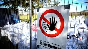Warnschild auf einem Zaun mit dem Hinweis (Asbestfasern). Dahinter liegen etliche weiße Säcke