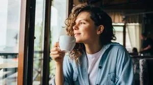 Smiling calm young woman drinking coffee looking out the window sitting at table in restaurant.