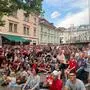 Mehrere Menschen stehen und sitzen auf einem Platz, manche tragen Fußballtrikots