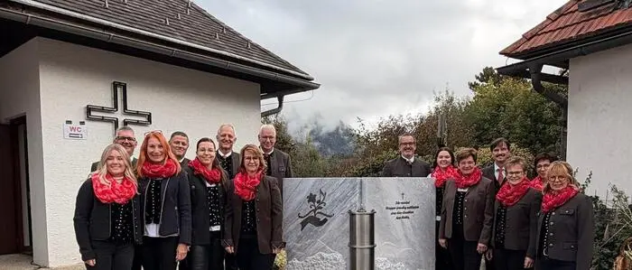 Mehrere Männer und Frauen stehen um einen Grabstein. Sie tragen einheitliche Kleidung, im linken Hintergrund sieht man ein Kreuz an der Hauswand