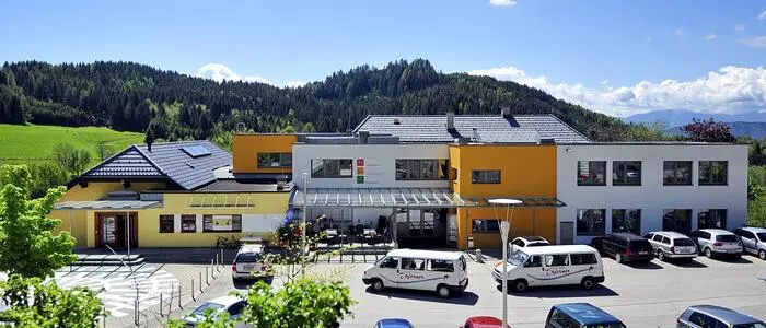 Zu sehen ist ein Parkplatz, dahinter ein Gebäudekomplex, in dem auch der Kindergarten untergebracht ist.