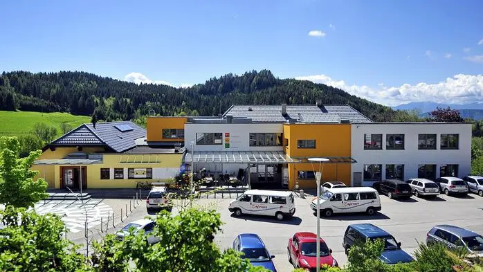 Zu sehen ist ein Parkplatz, dahinter ein Gebäudekomplex, in dem auch der Kindergarten untergebracht ist.