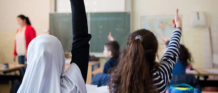 Das Bild zeigt zwei Mädchen, die in einem Klassenzimmer sitzen und aufzeigen. Eines der Mädchen trägt ein Kopftuch