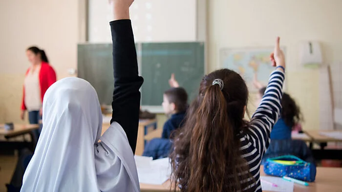 Das Bild zeigt zwei Mädchen, die in einem Klassenzimmer sitzen und aufzeigen. Eines der Mädchen trägt ein Kopftuch