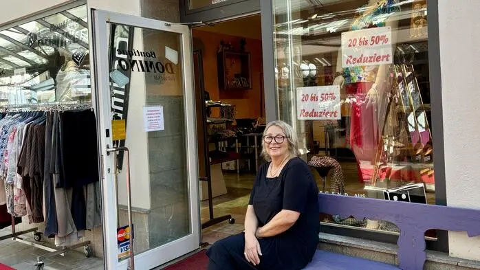 Eine Frau mit Brille sitzt vor einer offenen Geschäftstür auf einer lila Bank