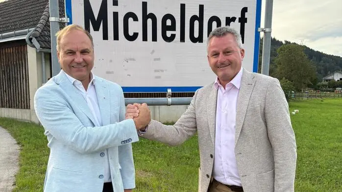 Zwei Männer stehen vor der Ortstafel in Micheldorf, geben sich die Hand und lächeln in die Kamera.