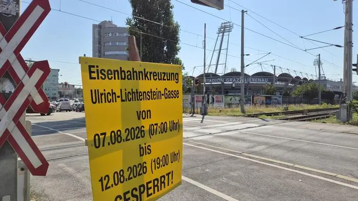 Baustellenhinweis direkt vor der Eisenbahnkreuzung mit der Aufschrift „Eisenbahnkreuzung Ulrich-Lichtenstein-Gasse von 07.08.2026 (07:00 Uhr) bis 12.08.2026 (19:00 Uhr) GESPERRT!“; im Hintergrund das Stadion Graz-Liebenau mit Stadionturm und Flutlichtmasten