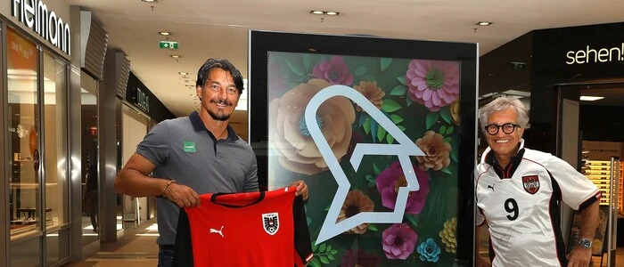 Zwei Männer stehen neben einer digitalen Anzeigetafel, einer trägt ein weißes Trikot der österreichischen Fußball-Nationalmannschaft, der andere hält ein rotes Trikot in seinen Händen.