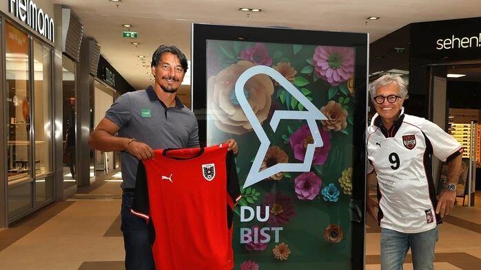 Zwei Männer stehen neben einer digitalen Anzeigetafel, einer trägt ein weißes Trikot der österreichischen Fußball-Nationalmannschaft, der andere hält ein rotes Trikot in seinen Händen.