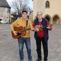 Zwei Männer, einer mit Gitarre und der andere mit einer Ukulele in der Hand, stehen auf einem großen Platz, im Hintergrund ist eine Kirche