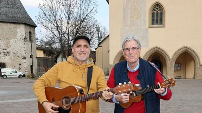 Zwei Männer, einer mit Gitarre und der andere mit einer Ukulele in der Hand, stehen auf einem großen Platz, im Hintergrund ist eine Kirche