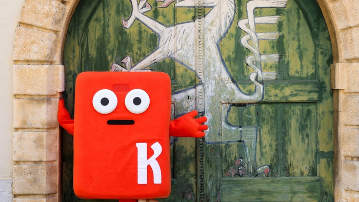 Bild zeigt Koini im Grazer Landhaus, er ist orange und kastig mit kurzen Händen und Füßen und einem K am Körper