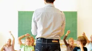 Zu sehen ist ein Lehrer der vor einer Klasse steht, mit dem Rücken zur Kamera, die Kinder im Hintergrund heben die Hand