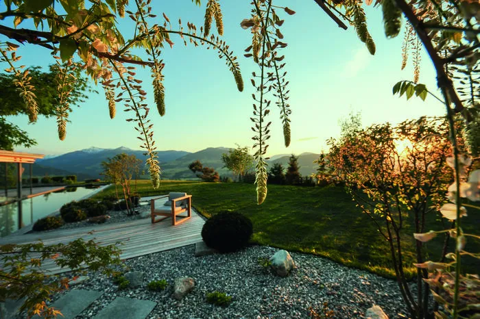 Die große Holzterrasse, die von Glas- wänden umgeben vor dem Wetter schützt, mit Stauden bepflanzte Kiesflächen und der Pool gehen fließend in den naturnahen Garten über. Ein Höhepunkt im Jahreslauf ist die Blüte der weißen Glyzinie, die die Pergola berankt, ein weiterer das Verfärben der Gruppe mächtiger Ahorn- bäume im Herbst.