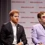 Prinz Harry sitzt in einem schwarzen Anzug neben Elton John, der einen lavendelfarbenen Anzug trägt.
