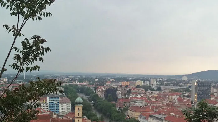Die Mur und ihr grünes Band - die dicht bewachsenen Uferböschungen auf beiden Seiten. Im Vordergrund glänzt das silbenere Kastner-Dach, dahinter ragt die Franziskanerkirche heraus.