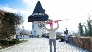 Das Bild zeigt Lukas Schönberger vor dem Uhrturm am Grazer Schlossberg mit den Langlaufski, einer pinken Haube und mit seiner pinken Unterhose in der Hand